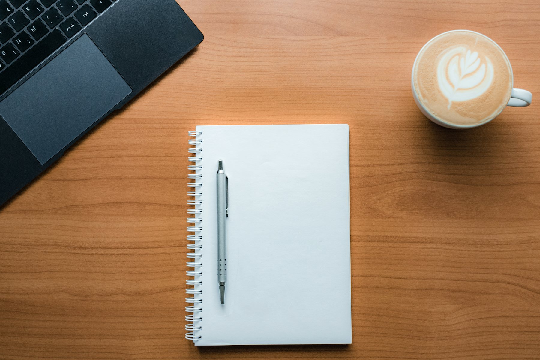 A notebook, pen and coffee beside a laptop, seen from above